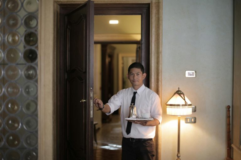 Friendly ethnic male waiter serving clients in room and carrying tray with coffee while working in stylish hotel